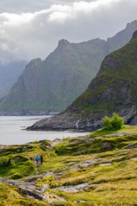 Two hikers on a grassy, rocky trail overlooking a bay with dramatic, cloud-covered mountains in the background under a cloudy sky.